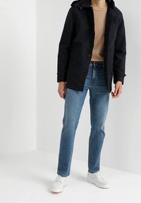 Schwarze wasserdichte Jacke mit Knopfverschluss und Kapuze, kombiniert mit hellblauen Jeans und weißen Turnschuhen. Einfaches Design und moderne Passform.