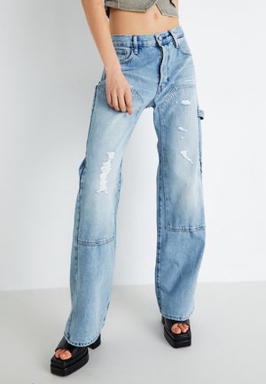 Frau trägt hellblaue, ausgefranste Jeans mit weitem Bein, Patches und schwarzen Plateau-Sandalen mit offenem Zehenbereich vor einfachem Hintergrund.