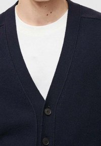 Námořnicky modrý cardigan s výstřihem do V, vyrobený z pletené látky, s dvěma tmavými knoflíky a nošený přes bílou košili.