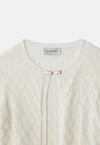 Cardigan en maille texturée blanche avec encolure ronde, deux boutons-pression décoratifs dorés et blancs, et étiquette "ANNA FIELD" à l'intérieur du col.