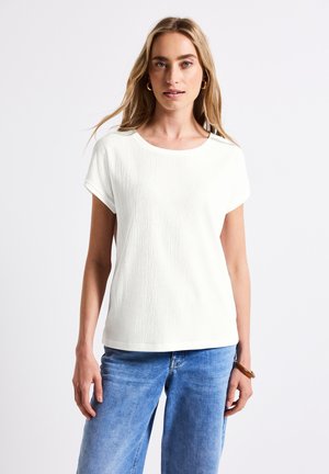 Witte, gestructureerde t-shirt met korte mouwen en een ronde halslijn, gecombineerd met lichtblauwe jeans. De stof heeft een subtiele, gekreukte afwerking.