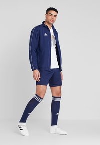 Marineblaue Sportjacke mit weißen Streifen, kombiniert mit einem weißen Shirt, marineblauen Shorts und knielangen marineblauen Socken mit drei weißen Streifen.