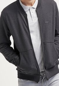 Homme portant une veste zippée gris foncé par-dessus un polo gris clair, les mains dans les poches de la veste.