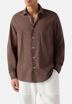 Uomo che indossa una camicia marrone a maniche lunghe con bottoni bianchi e pantaloni bianchi, mani nelle tasche, di fronte.