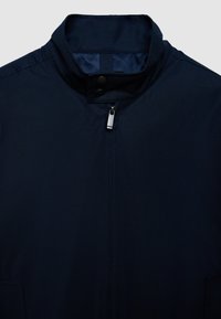 Giacca blu navy con colletto alto, dotata di chiusura con zip e due bottoni a pressione. Tessuto liscio, senza motivi visibili, design minimalista.