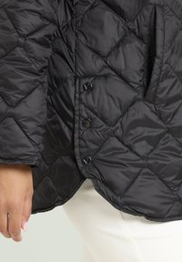 Giubbotto imbottito nero con cuciture a rombi, tre bottoni a pressione e una tasca laterale, indossato sopra pantaloni bianchi con una mano visibile.