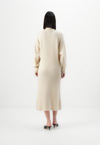 Vestito in maglia beige con maniche lunghe, di lunghezza media, texture a costine e spacchi laterali; abbinato a scarpe con tacco alto nere.