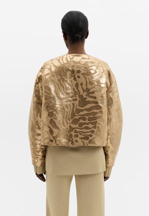 Personne aux cheveux foncés en chignon bas portant une veste dorée brillante et à motif sur des vêtements beiges superposés, vue de dos sur un fond blanc uni.