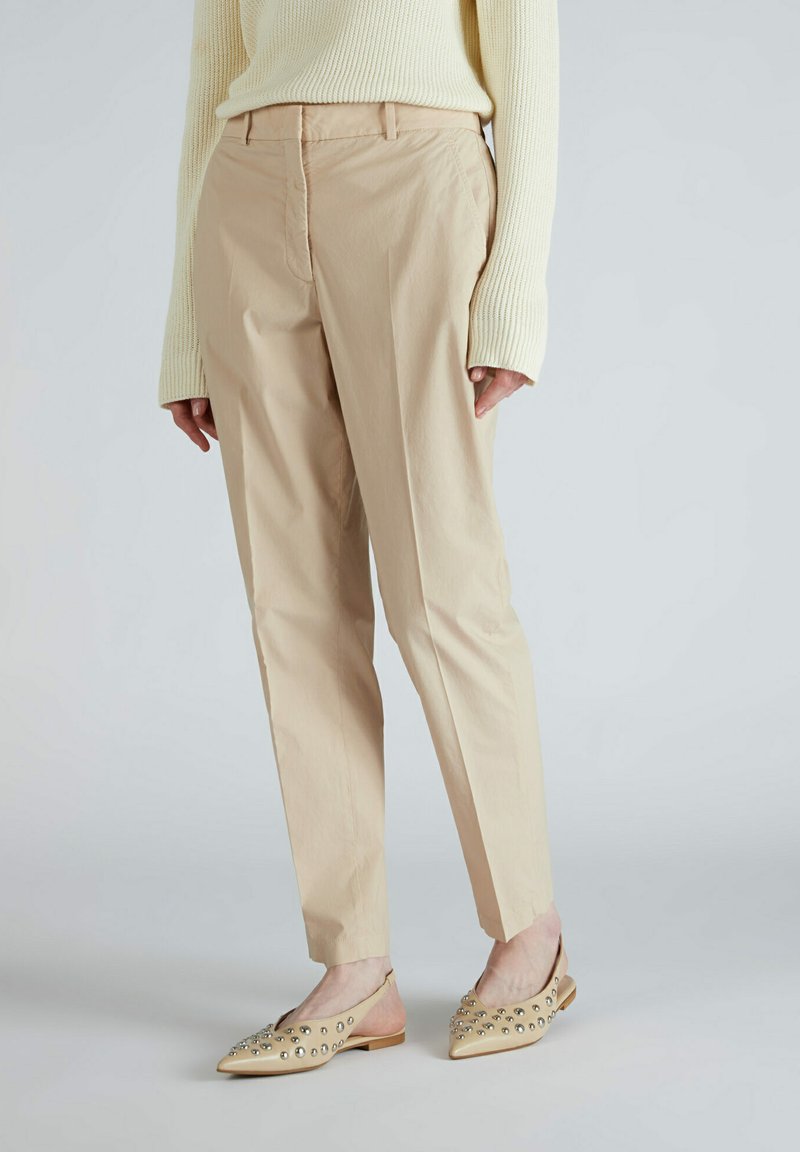 Pantalons en coton beige avec une coupe ajustée, présentant un devant plat et des poches latérales. Portés avec des chaussures plates beige ornées de clous.