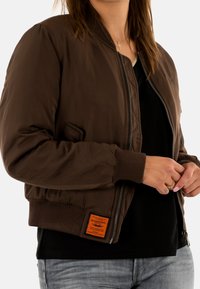 Giacca bomber marrone con zip frontale, polsini e orlo a coste, e un'etichetta arancione. Indossata sopra una camicia nera, con una texture liscia.