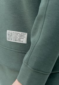 Zelená sweatshirt s neoprenovým efektom, vyrobená z pletenej tkaniny, s 4-smerným elastickým materiálom a etiketou, ktorá uvádza elastické, teplé, rýchlo schnúce a antibakteriálne vlastnosti.