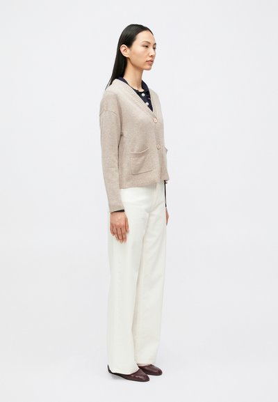 Γυναίκα με μακριά μαύρα μαλλιά, φορίζοντας μια μπεζ πλεκτή ζακέτα με κουμπιά, λευκό παντελόνι φαρδιάς γραμμής, σκοτεινά παπούτσια, στέκεται μπροστά σε ένα απλό ανοιχτόχρωμο φόντο.