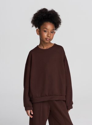 Felpa marrone oversize con maniche lunghe e scollo rotondo, realizzata in un tessuto morbido e liscio. Caratterizzata da una vestibilità comoda e polsini a coste.