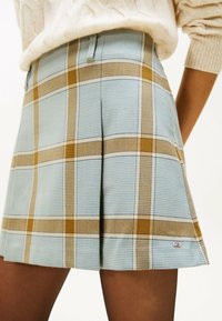 Vrouw in een blauw-beige geruite geplooide rok, gecombineerd met een crèmekleurige gebreide trui, die tot halverwege de dij komt en een klein logo-embleem toont.