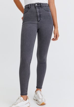 Graue Slim-Fit Jeans mit hoher Taille, getragen mit weißen Sneakers an einer Person mit weißem Oberteil, stehend vor einem einfarbigen hellen Hintergrund.
