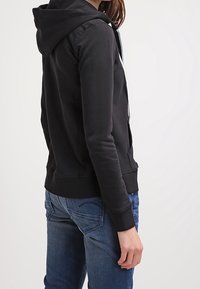 Felpa nera con zip e cappuccio, con polsini e orlo a coste. Indossata con jeans in denim blu. Tessuto liscio, design minimalista senza motivi.