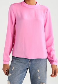 Blusa rosa a maniche lunghe con scollo a girocollo, realizzata in tessuto morbido. Presenta polsini elasticizzati e una vestibilità comoda, abbinata a jeans blu strappati.