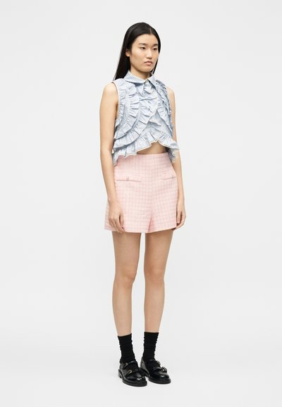 Νέα γυναίκα με μακριά σκουρόχρωμα μαλλιά, ντυμένη με  μπλε ριχτό crop τοπ χωρίς μανίκια, ροζ υφασμάτινη βερμούδα, μαύρες κάλτσες και μαύρα μοκασίνια, στέκεται ακίνητη.