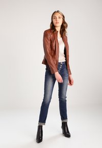 Brun læderjakke med syede skulderdetaljer, båret over en hvid top, sammen med tætsiddende blå jeans og sorte ankelstøvler.