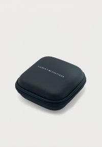 Στενό συμπαγές σκληρό θήκη σκούρου μπλε χρώματος με φερμουάρ, με το λογότυπο Tommy Hilfiger στο κέντρο στο πάνω μέρος, τοποθετημένη σε απλό ανοιχτόχρωμο φόντο.