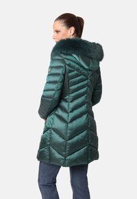 Mujer que lleva un abrigo de invierno acolchado en color verde azulado con capucha forrada de piel, de pie de lado con las manos en los bolsillos, combinado con unos vaqueros azules.