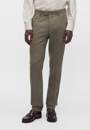 Homme portant un pantalon marron sur mesure, une chemise blanche et des mocassins en cuir marron foncé, debout devant un fond uni.