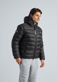 Sort puffer-jakke med lynlåslukning foran, høj krave og hætte. Har quiltet tekstur og logo-detaljer på brystet og ærmet.