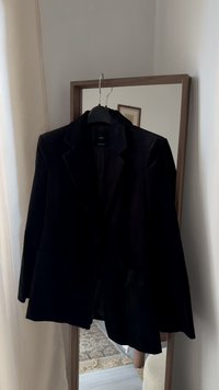 Czarna aksamitna marynarka zawieszona na wieszaku, z klapą z wycięciem, zapinana na jeden guzik, z dwoma przednimi kieszeniami. Materiał wydaje się miękki.