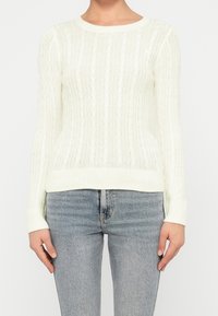Personne portant un pull en maille torsadée blanc et un jean bleu clair, debout avec les bras détendus le long du corps.