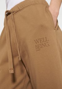 Bruine katoenen sweatpants met een koord, voorzien van een subtiele "WELL BEING." print op de zijkant. Glad materiaal en een relaxed fit.