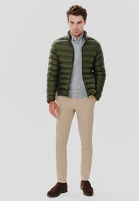 Oliwkowa kurtka puchowa z poziomym przeszyciem, szary sweter pod spodem, beżowe spodnie slim-fit i ciemnobrązowe buty wsuwane.