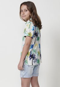 Camisa de manga corta de tela blanca con un colorido patrón de palmeras en verde, azul y negro, combinada con pantalones cortos de mezclilla claros.