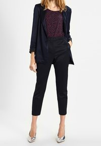 Mujer vestida con una chaqueta azul marino, blusa de patrón burdeos, pantalones negros hasta los tobillos y tacones altos plateados de punta, de pie con una mano en el bolsillo.