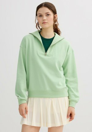 Joven con cabello castaño que lleva una sudadera verde claro con media cremallera sobre una camiseta verde oscuro y una falda blanca plisada, posando frente a un fondo liso.