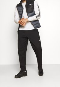 Schwarze Steppweste mit grauen Akzenten über einem weißen Langarmshirt, kombiniert mit schwarzen Hosen und schwarzen Sportschuhen. North Face-Logo sichtbar.