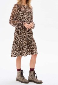 Robe à imprimé léopard avec un col à volants et des manches longues, présentant un ourlet en étages. Associée à des bottes à lacets assorties au motif léopard.