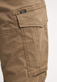 Pantalones cortos de cargo color caqui con un bolsillo de solapa, costuras visibles y una pequeña etiqueta de marca azul. La tela tiene una textura suave y mate.