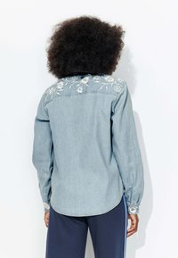 Chemise en denim bleu clair avec des broderies florales sur le dos, manches longues, poignets boutonnés et ourlet incurvé.