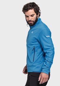 Blaue Zip-Jacke mit strukturierter Oberfläche, ausgestattet mit einem hohen Kragen und einer seitlichen Zippertasche. Akzente umfassen ein Logo auf dem Ärmel.