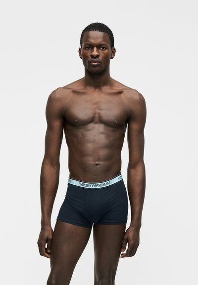 Μαύρος άνδρας μοντέλο στέκεται, φορώντας μαύρα αντρικά εσώρουχα Emporio Armani με ανοιχτό μπλε ζώνη μέσης, μπροστά σε απλό ανοιχτό γκρι φόντο.