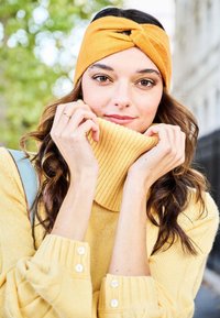 Fascia per capelli gialla lavorata a maglia con un design a spirale, abbinata a un maglione giallo a collo alto con coste e polsini con bottoni.