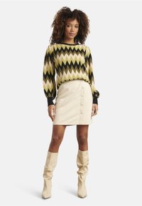 Multicolored zigzag-patterned sweater in olive, black, and cream, paired with a beige faux suede A-line skirt featuring front buttons and knee-high boots.