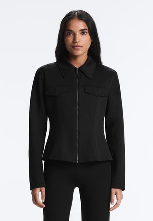 Žena v priliehavej čiernej bunde so zipsom vpredu, širokým golierom a dvoma náprsnými vreckami so záklopkou, stojaca proti jednoliatemu pozadiu.