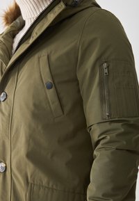 Chaqueta de invierno verde oliva con cierre frontal de botones y presión, bolsillo lateral en el pecho, bolsillo con cremallera en la manga, usada sobre un suéter de punto color crema.