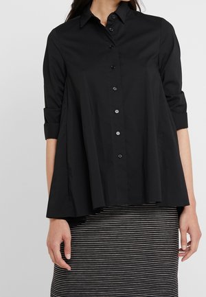 Femme portant une chemise noire ample boutonnée avec les manches retroussées et une jupe rayée horizontale noire et blanche.