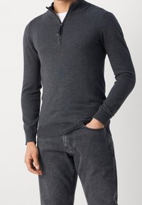 Maglione grigio scuro con zip al collo in un tessuto morbido, caratterizzato da maniche lunghe, polsini e orlo a coste e una trama liscia. Abbinato a jeans scuri.