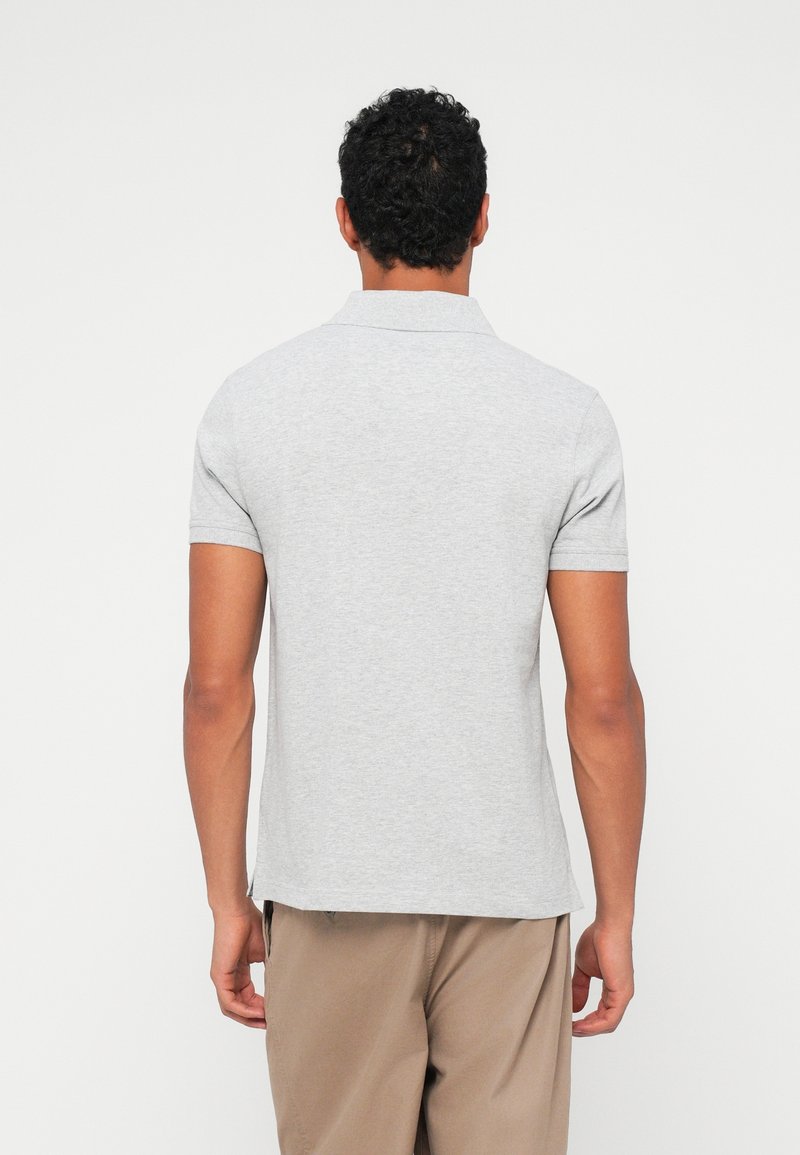 Homme aux cheveux courts et bouclés, portant un polo gris clair à manches courtes et un pantalon beige, debout de dos sur fond blanc.