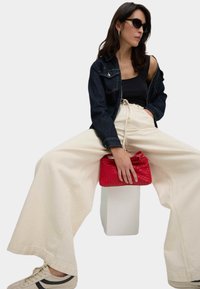 Weite beige Hosen mit einem Kordelzug in der Taille, ein schwarzes Tanktop und eine dunkelblaue Jeansjacke. Hält eine rote, gewebte Handtasche und trägt schwarze Sonnenbrillen.