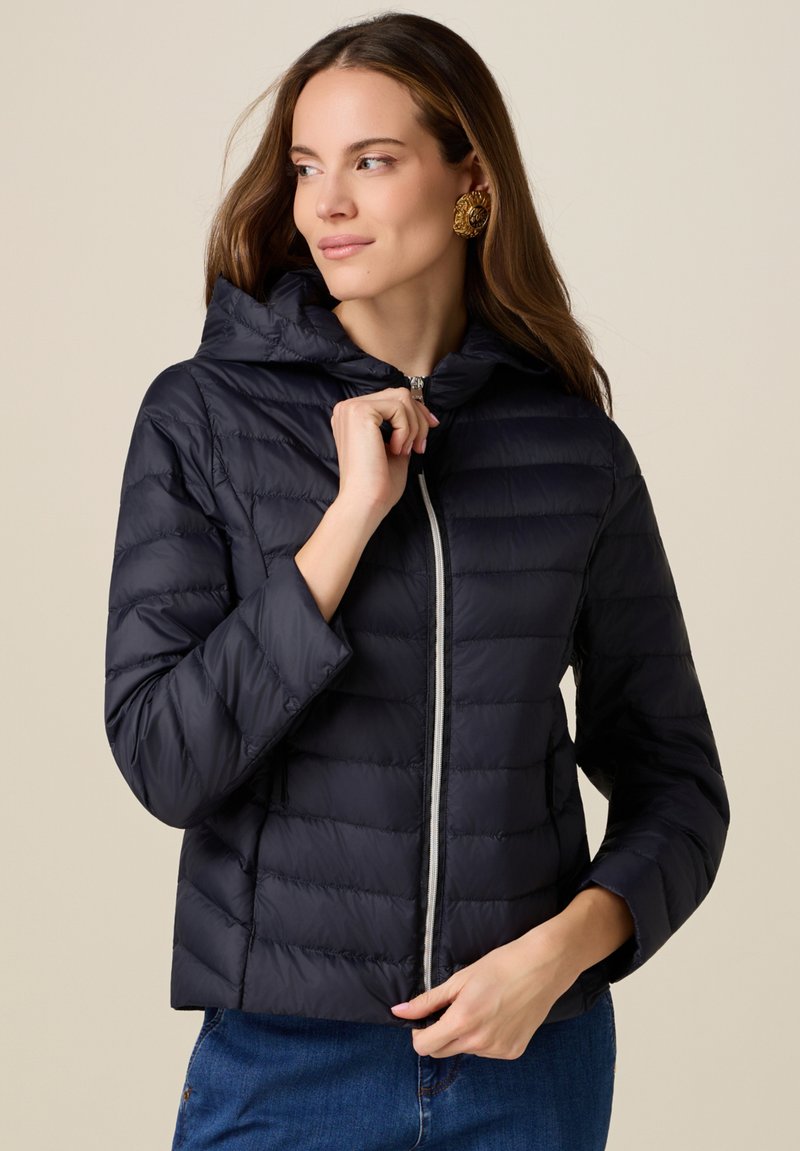 Frau trägt eine dunkle gesteppte Jacke mit Kapuze, schließt den Reißverschluss und trägt eine blaue Jeans vor einem schlichten beigen Hintergrund.