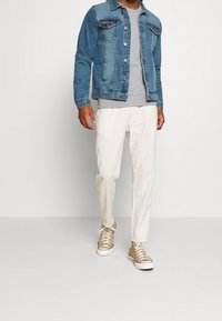 Giacca di jeans blu con bottoni metallici, t-shirt grigia, pantaloni bianchi a righe e sneakers marroni chiari con suole bianche.